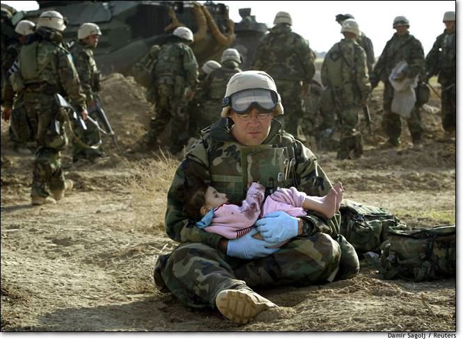 A soldier dressed in camo sitting on dirt holding a little girl in pink on his lap